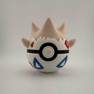 Może przedstawiać: Biało-czarna Pokeball w stylu Togepi z czerwonymi i niebieskimi akcentami. G&oacute;rna część jest w jasnobrązowym kolorze z kilkoma spiczastymi wypustkami. Projekt obejmuje czarny pasek i centralny czarny okrąg.