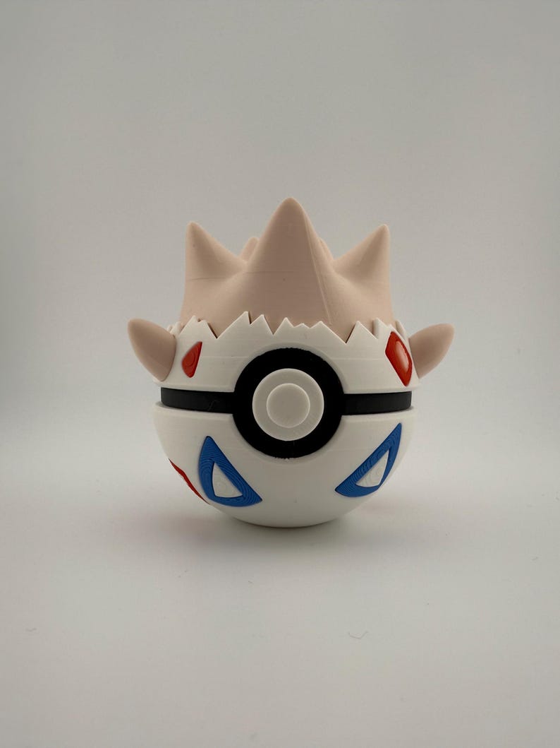 Może przedstawiać: Biało-czarna Pokeball w stylu Togepi z czerwonymi i niebieskimi akcentami. G&oacute;rna część jest w jasnobrązowym kolorze z kilkoma spiczastymi wypustkami. Projekt obejmuje czarny pasek i centralny czarny okrąg.