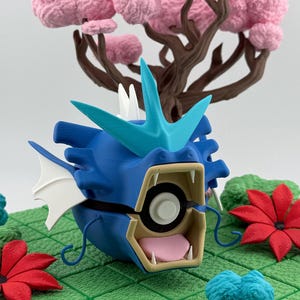 May include: A blue and turquoise 3D-printed figure of a dragon-like creature with an open mouth, white teeth, and a pink tongue. The figure is set on a green surface with red and blue floral accents and a pink tree in the background.