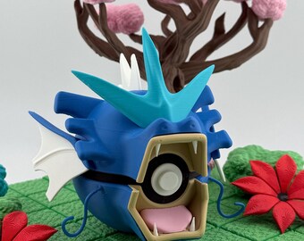 Op de afbeelding: Een blauw en turquoise 3D-geprint figuur van een draakachtig wezen met een open mond, witte tanden en een roze tong. Het figuur staat op een groene ondergrond met rode en blauwe bloemaccenten en een roze boom op de achtergrond.