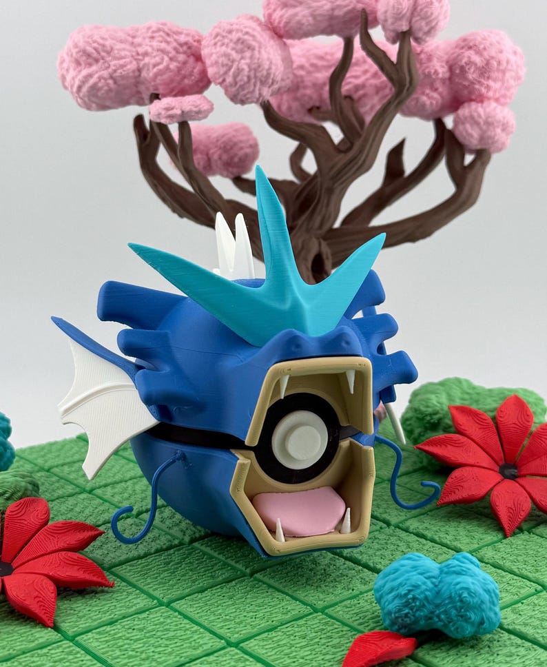 May include: A blue and turquoise 3D-printed figure of a dragon-like creature with an open mouth, white teeth, and a pink tongue. The figure is set on a green surface with red and blue floral accents and a pink tree in the background.