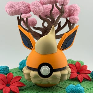 May include: A 3D-printed orange and tan figure with black accents, resembling a character from a popular animated series. It is set against a backdrop of a pink tree and green, textured ground with red and blue floral accents.