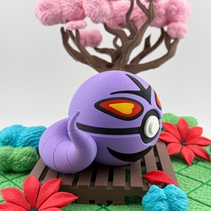 May include: A purple, spherical creature with black and yellow accents, a curved tail, and a white eye, resting on a brown wooden platform. The scene includes a pink tree and red and blue floral elements, set against a green, textured background.