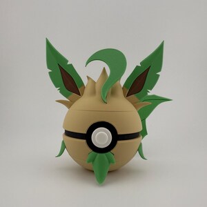 May include: A tan and green ball-shaped ornament with a black stripe and a white circle in the centre. It features green leaf-like structures and brown accents, resembling a stylized plant or creature.