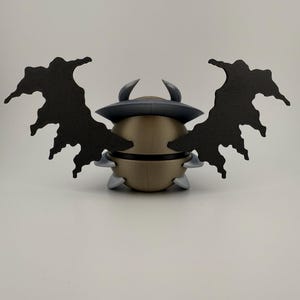 May include: A gold-colored, spherical figurine with a dark gray hat and two curved horns. The figurine has two large, black, wing-like attachments. Small, gray, triangular shapes are attached to the base of the figurine.