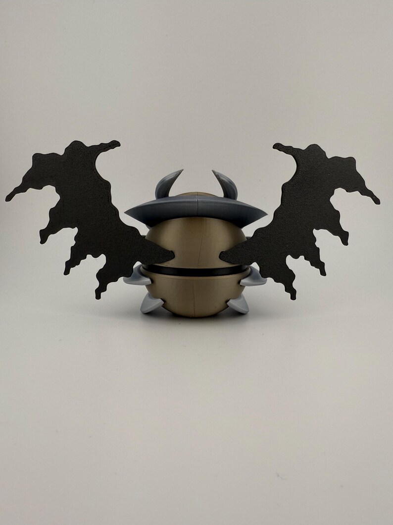 May include: A gold-colored, spherical figurine with a dark gray hat and two curved horns. The figurine has two large, black, wing-like attachments. Small, gray, triangular shapes are attached to the base of the figurine.