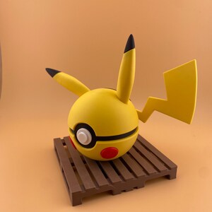 May include: A yellow Pikachu-themed sphere with black-tipped ears and a lightning bolt tail. The sphere has a black stripe, white circle, and red accents. It rests on a brown wooden pallet, set against a solid orange background.