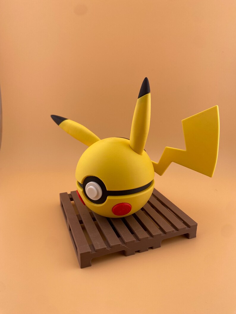 May include: A yellow Pikachu-themed sphere with black-tipped ears and a lightning bolt tail. The sphere has a black stripe, white circle, and red accents. It rests on a brown wooden pallet, set against a solid orange background.