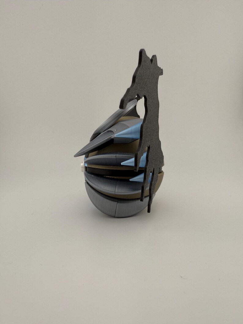 May include: A multi-layered, abstract sculpture with a rounded base and a jagged, dark gray silhouette extending upwards. The sculpture features shades of gray, blue, and brown, creating a unique, modern design. The overall shape is reminiscent of a stylized grenade.