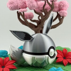 May include: A decorative silver and white sphere with rabbit ears and a tail, set against a backdrop of a pink-flowered tree and red flowers. The scene is on a green, grid-patterned surface.