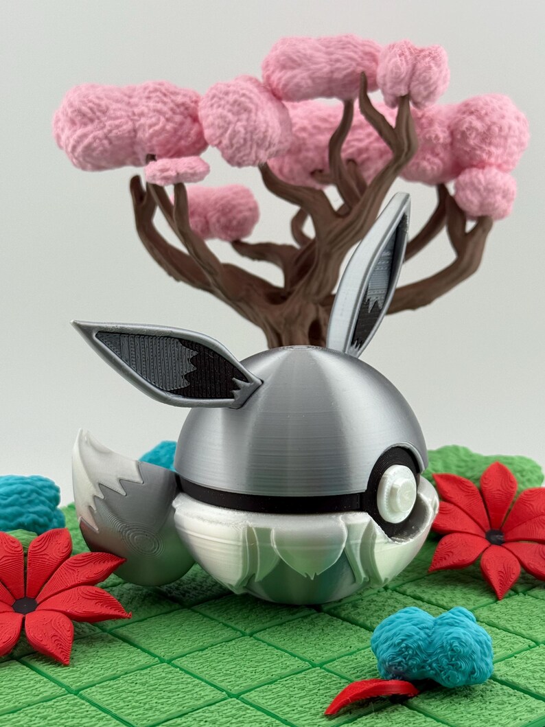 May include: A decorative silver and white sphere with rabbit ears and a tail, set against a backdrop of a pink-flowered tree and red flowers. The scene is on a green, grid-patterned surface.