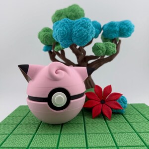 K&ouml;nnte beinhalten: Ein rosa-schwarzer Pok&eacute;ball im Jigglypuff-Design liegt auf einer gr&uuml;nen, gefliesten Oberfl&auml;che. Eine rote Blume und ein Miniaturbaum mit blauem und gr&uuml;nem Laub befinden sich im Hintergrund. Der Pok&eacute;ball hat einen schwarzen Streifen und einen wei&szlig;en Knopf.