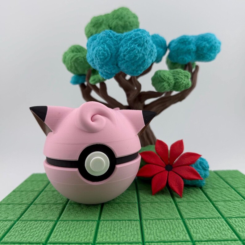 K&ouml;nnte beinhalten: Ein rosa-schwarzer Pok&eacute;ball im Jigglypuff-Design liegt auf einer gr&uuml;nen, gefliesten Oberfl&auml;che. Eine rote Blume und ein Miniaturbaum mit blauem und gr&uuml;nem Laub befinden sich im Hintergrund. Der Pok&eacute;ball hat einen schwarzen Streifen und einen wei&szlig;en Knopf.