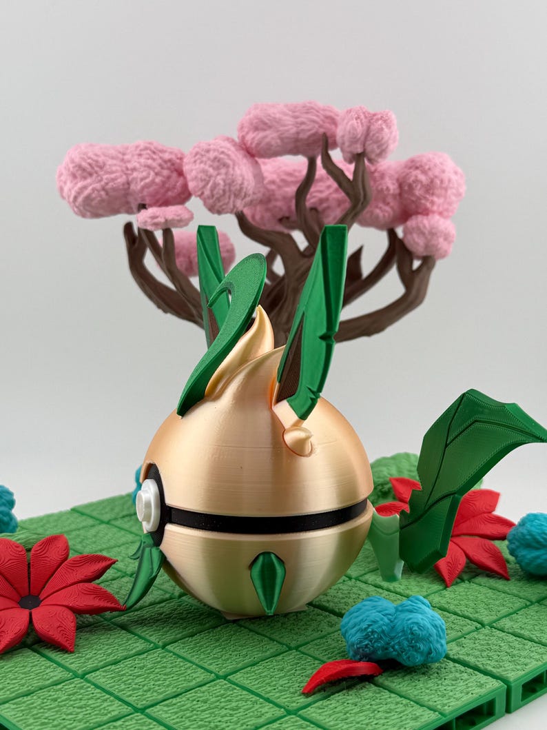 May include: A gold-coloured, spherical object with green leaf-like accents and a black stripe, set on a green grid. A pink tree with brown branches and pink fluffy foliage is in the background. Red and blue floral accents are scattered around the base.