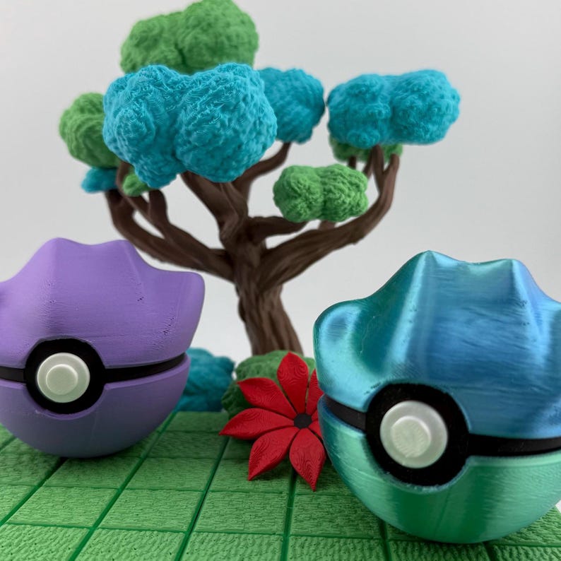 May include: A diorama featuring two colorful, stylized ball-shaped objects, one purple and one teal, with white accents. A miniature tree with green and blue foliage and a red flower are in the background, set on a green, grid-patterned base.