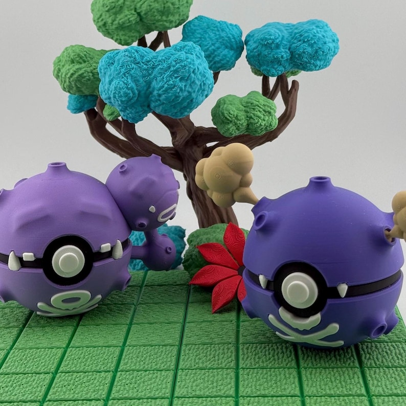 May include: A diorama featuring two purple cartoon characters with white accents, set against a backdrop of a miniature tree with green and blue foliage. A red leaf and green base complete the scene.