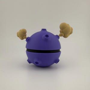 May include: A purple, spherical 3D-printed object with a black band around the middle and tan cloud-like shapes extending from the sides. The object has several circular openings and a small cylindrical protrusion on top.
