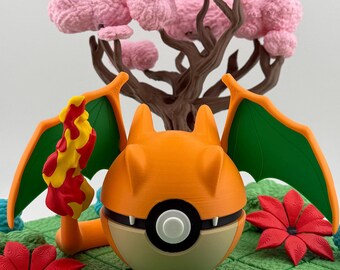 Op de afbeelding: Een oranje en bruin Charizard-thema Poké Ball figuur met groene vleugels en een vlam. De figuur staat op een groene, rasterpatroon basis met rode en blauwe bloemaccenten en een roze boom. Een uniek verzamelobject.