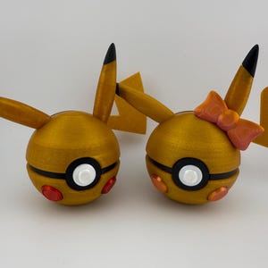 May include: Two golden-yellow, spherical figures with black and white details, resembling cartoon characters. One has a bow. Each has black-tipped ears and red and orange accents. The figures are likely toys or decorative items.