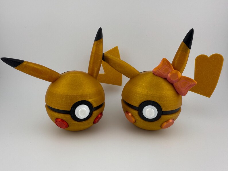 May include: Two golden-yellow, spherical figures with black and white details, resembling cartoon characters. One has a bow. Each has black-tipped ears and red and orange accents. The figures are likely toys or decorative items.