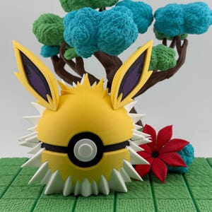 May include: A yellow and white 3D-printed Jolteon-themed Poke Ball with spiky details and black accents. The design includes yellow ears with purple interiors. A miniature tree with green and blue foliage is in the background.