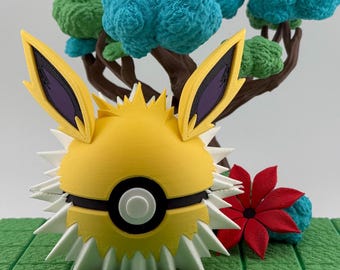 Op de afbeelding: Een geel en witte 3D-geprinte Jolteon-thema Poké Ball met stekelige details en zwarte accenten. Het ontwerp bevat gele oren met paarse interieurs. Een miniatuurboom met groene en blauwe bladeren staat op de achtergrond.
