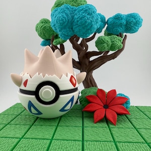 Może przedstawiać: Wydrukowana w 3D figurka Togepi z białym i czarnym korpusem, czerwonymi akcentami i niebieskimi tr&oacute;jkątami. Za nią stoi miniaturowe drzewo z niebieskimi i zielonymi liśćmi, a obok czerwony kwiat na zielonej, teksturowanej podstawie.