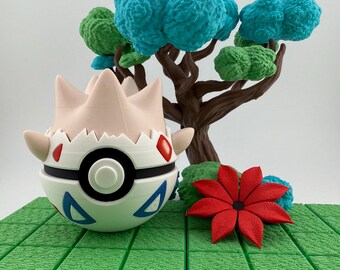 Op de afbeelding: Een 3D-geprinte Togepi-figuur met een wit en zwart lichaam, rode accenten en blauwe driehoeken. Een miniatuurboom met blauw en groen gebladerte staat erachter, samen met een rode bloem op een groene, getextureerde basis.