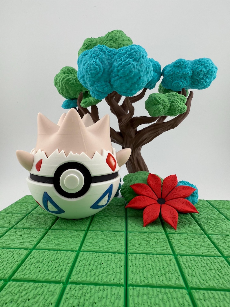 Może przedstawiać: Wydrukowana w 3D figurka Togepi z białym i czarnym korpusem, czerwonymi akcentami i niebieskimi tr&oacute;jkątami. Za nią stoi miniaturowe drzewo z niebieskimi i zielonymi liśćmi, a obok czerwony kwiat na zielonej, teksturowanej podstawie.