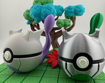 Op de afbeelding: Twee bolvormige, metaalachtig uitziende objecten met zwarte en witte accenten en groene en paarse details. Een paars, gebogen object en een rode bloem bevinden zich tussen de objecten. Een miniatuurboom met groen en blauw gebladerte staat op de achtergrond.