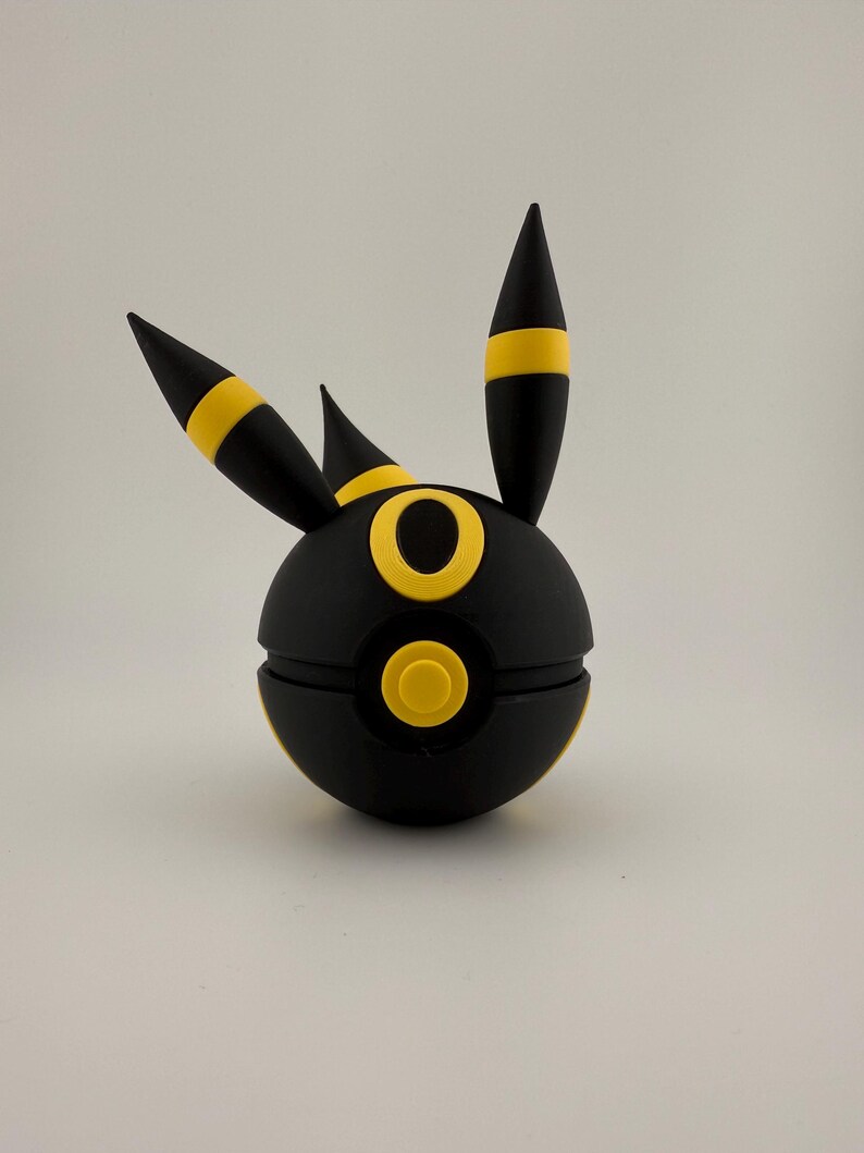 May include: A black and yellow spherical figure with three pointed projections. The figure has a circular yellow design on the front and yellow accents on the projections. The object is set against a plain white background.