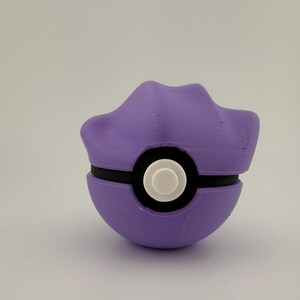 May include: A purple and black ball-shaped object with a white circular center. The top portion has a unique, curved design. The object is likely a toy or collectible item, possibly inspired by a popular animated series.