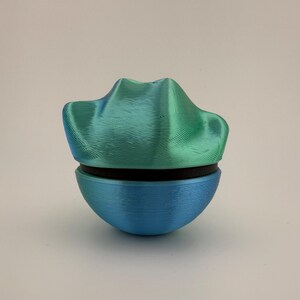 May include: A two-part, 3D-printed decorative object. The top section is a textured, teal-green color, resembling a stylized mountain range. The bottom section is a smooth, rounded shape in blue. A black band separates the two parts.