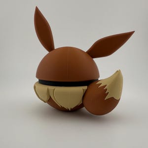 May include: A brown and tan Eevee figure, with large ears and a fluffy tail. The figure has a round body with a black band around the middle. The tail and the lower part of the figure are tan, while the rest is brown.
