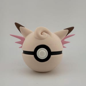 K&ouml;nnte beinhalten: Ein hellrosa und wei&szlig;er Pok&eacute;ball mit einem Cartoon-Charakter-Design. Die Figur hat braune Ohren, rosa Fl&uuml;gel und eine Wirbel auf der Oberseite. Der Ball hat einen schwarzen Streifen und einen wei&szlig;en Knopf.
