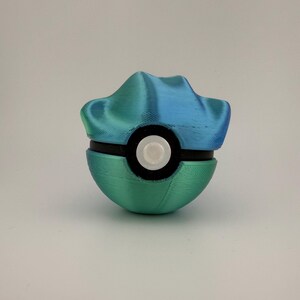 May include: A 3D-printed Pok&eacute; Ball in shades of teal and blue. The top section has a textured, mountain-like design. The ball is bisected by a black band with a white circular button in the center. The bottom half is a solid teal color.