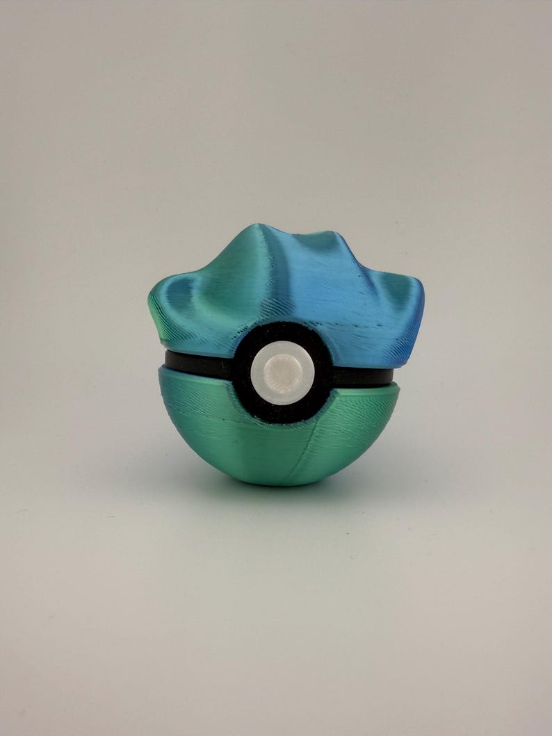 May include: A 3D-printed Pok&eacute; Ball in shades of teal and blue. The top section has a textured, mountain-like design. The ball is bisected by a black band with a white circular button in the center. The bottom half is a solid teal color.