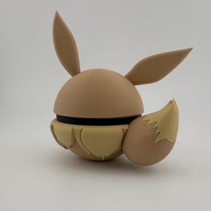 May include: A tan-coloured, 3D-printed figure of a cartoon character with large ears and a fluffy tail. The figure has a spherical body with a black band around the middle. The ears and tail are a lighter shade of tan.