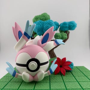 May include: A 3D-printed figurine of a Sylveon Pok&eacute;mon character, featuring a pink, white, and blue color scheme. The figure is set on a green textured base with a miniature tree and red flower. The Sylveon has large, ribbon-like appendages.
