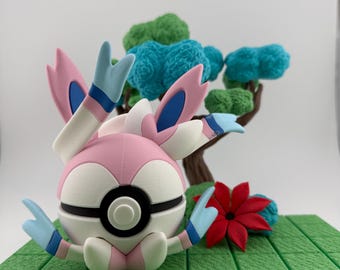 Op de afbeelding: 3D-geprinte figuur van een Sylveon Pokémon-personage, met roze, witte en blauwe kleuren. De figuur staat op een groene, getextureerde basis met een miniatuurboom en een rode bloem. Sylveon heeft grote, lintachtige aanhangsels.