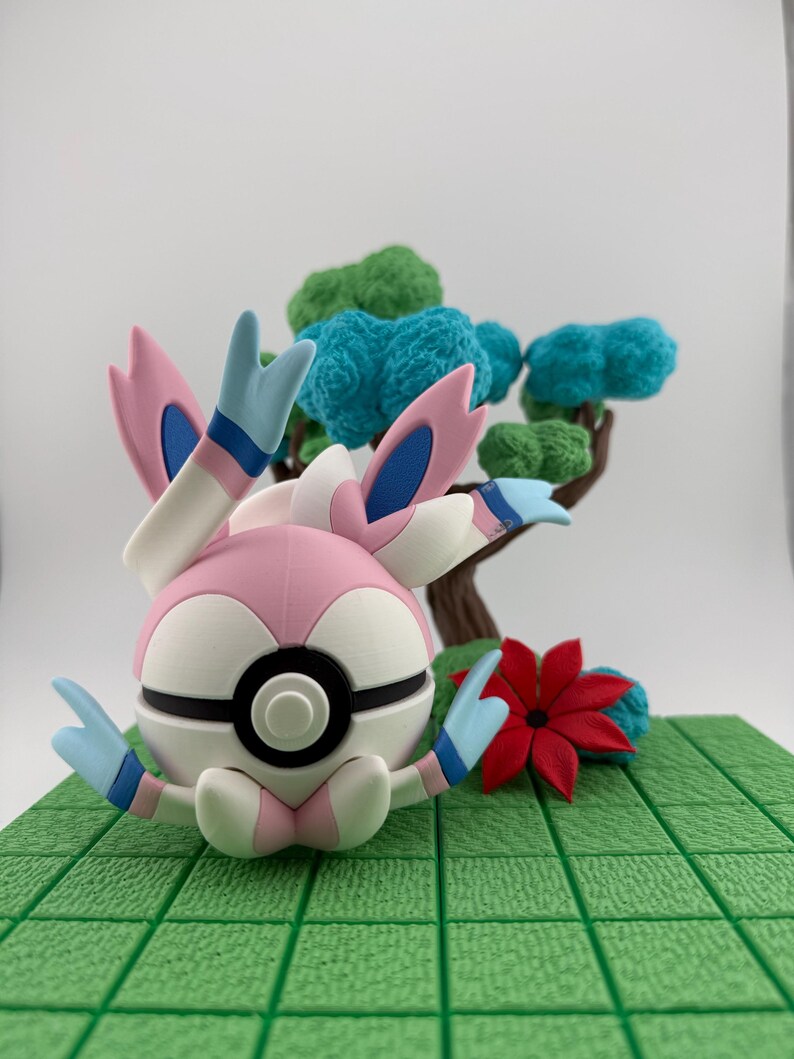 May include: A 3D-printed figurine of a Sylveon Pok&eacute;mon character, featuring a pink, white, and blue color scheme. The figure is set on a green textured base with a miniature tree and red flower. The Sylveon has large, ribbon-like appendages.