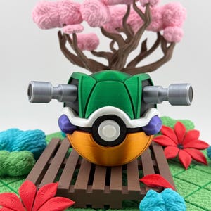 May include: A 3D-printed, stylized Poke Ball with a green shell, orange base, and silver side attachments. The ball rests on a brown platform with a miniature pink tree and red and blue floral accents in the background.