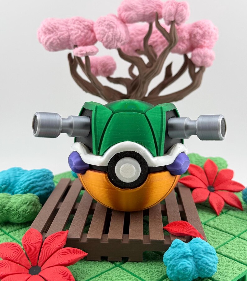 May include: A 3D-printed, stylized Poke Ball with a green shell, orange base, and silver side attachments. The ball rests on a brown platform with a miniature pink tree and red and blue floral accents in the background.