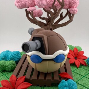 May include: A detailed sculpture of a cartoon character with a brown shell, a gray cannon, and blue eyes. The character is set in a miniature landscape with a pink tree, red flowers, and blue and green textured elements.