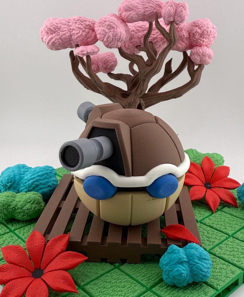 May include: A detailed sculpture of a cartoon character with a brown shell, a gray cannon, and blue eyes. The character is set in a miniature landscape with a pink tree, red flowers, and blue and green textured elements.