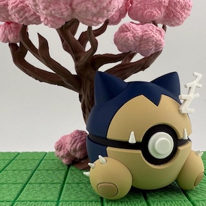 May include: A Snorlax figure in a sleeping pose, with a blue and tan color scheme, and white accents. The figure is set against a backdrop of a brown tree with pink blossoms and a green, grid-like base.