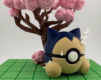 Op de afbeelding: Een Snorlax-figuur in slaaphouding, met een blauw en beige kleurenschema en witte accenten. De figuur staat tegen een achtergrond van een bruine boom met roze bloesems en een groene, rasterachtige basis.