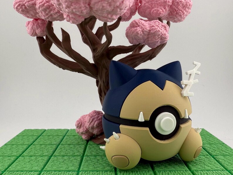 May include: A Snorlax figure in a sleeping pose, with a blue and tan color scheme, and white accents. The figure is set against a backdrop of a brown tree with pink blossoms and a green, grid-like base.