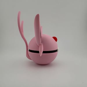 May include: A pink, spherical object with a black band around the middle, featuring a red dot and two pink ear-like projections. A curved pink tail extends from the back. The object is set against a white background.