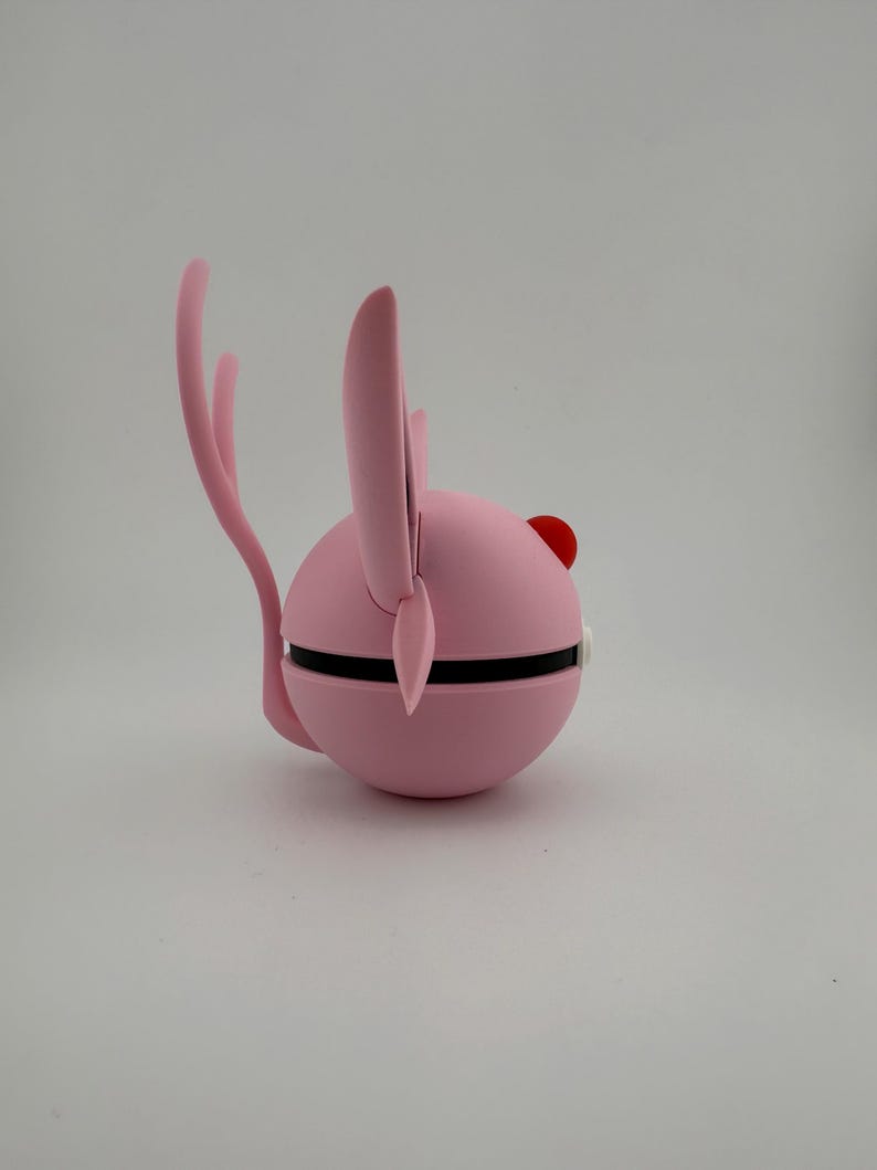 May include: A pink, spherical object with a black band around the middle, featuring a red dot and two pink ear-like projections. A curved pink tail extends from the back. The object is set against a white background.